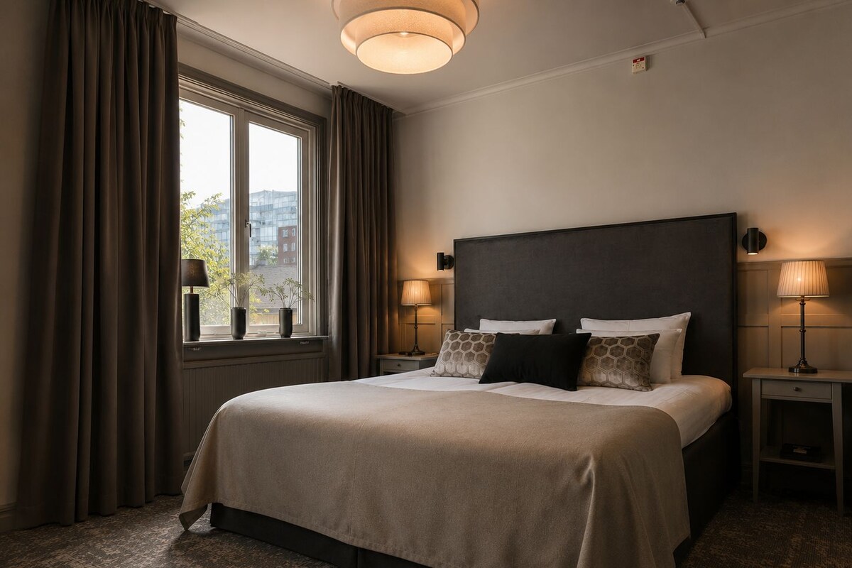 A hotel room with a large bed, two bedside tables, a window with curtains, and a view of buildings outside.