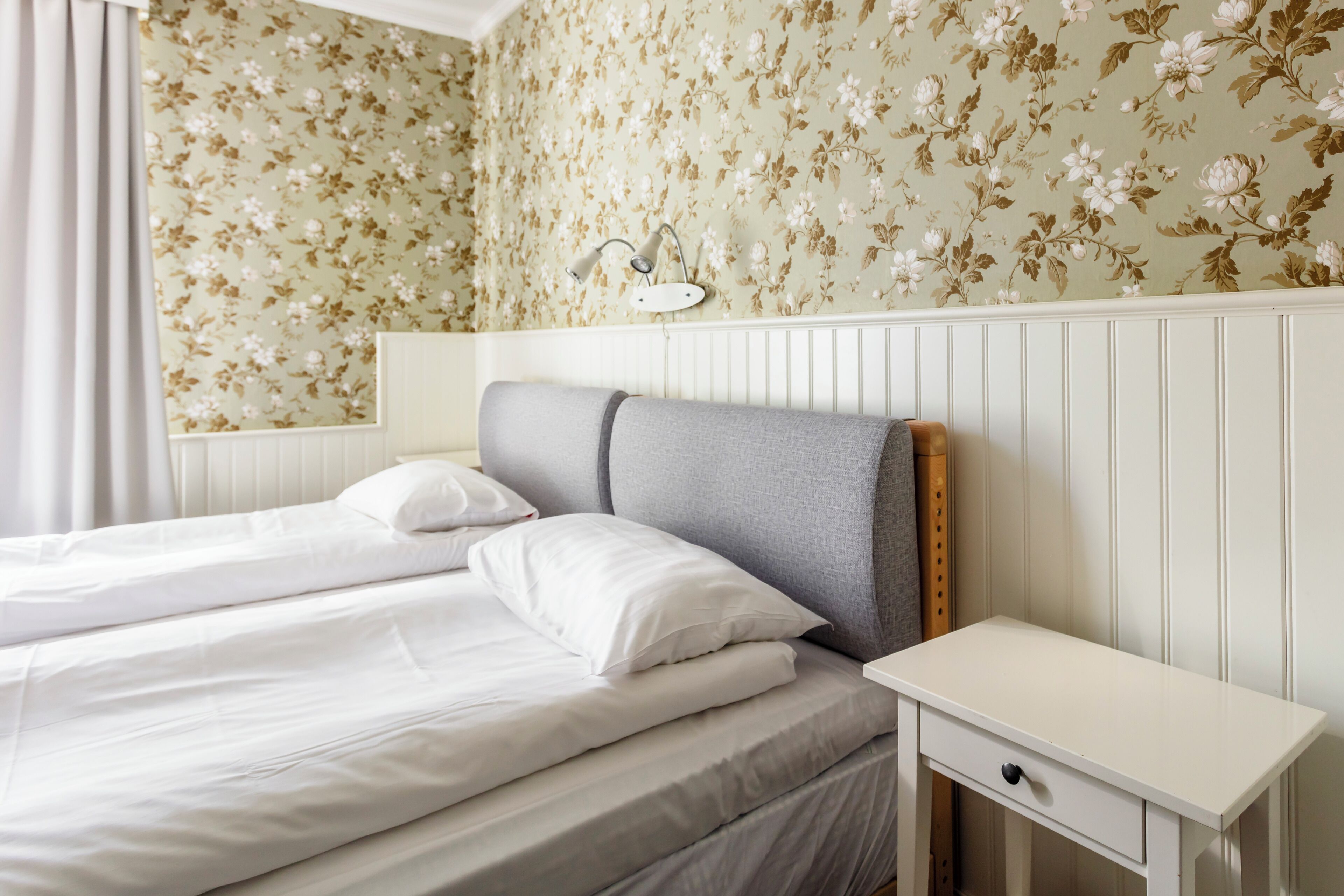 Una cama bien hecha con sábanas y almohadas blancas, una mesita de noche con cajón y una pared con papel tapiz floral.