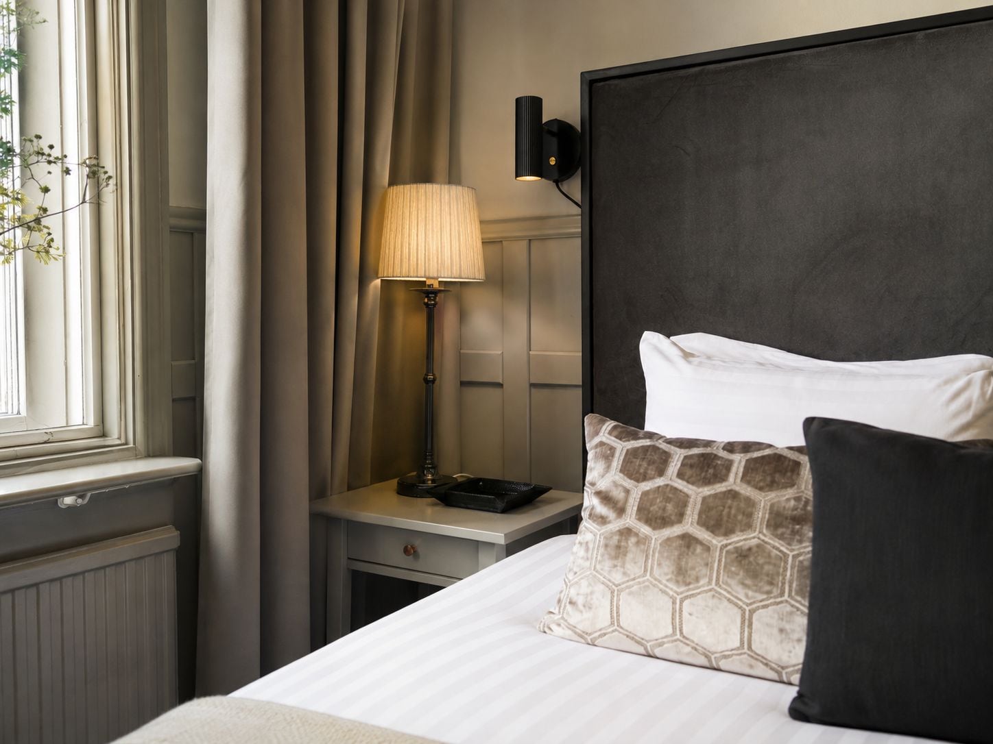 Ett hotellrum med en säng, ett sängbord, en lampa och ett fönster med gardiner.