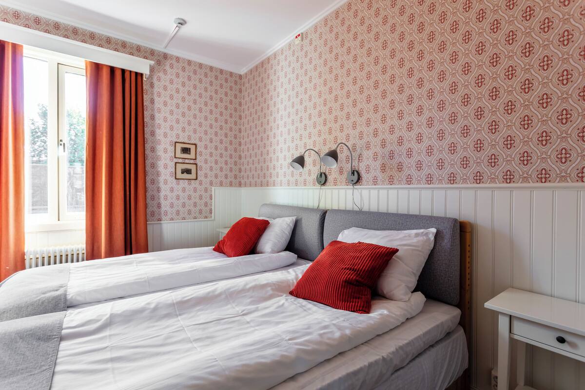 Ein Doppelbett mit weißer Bettwäsche und roten Kissen, ein Nachttisch mit Lampe sowie Tapete mit Muster.