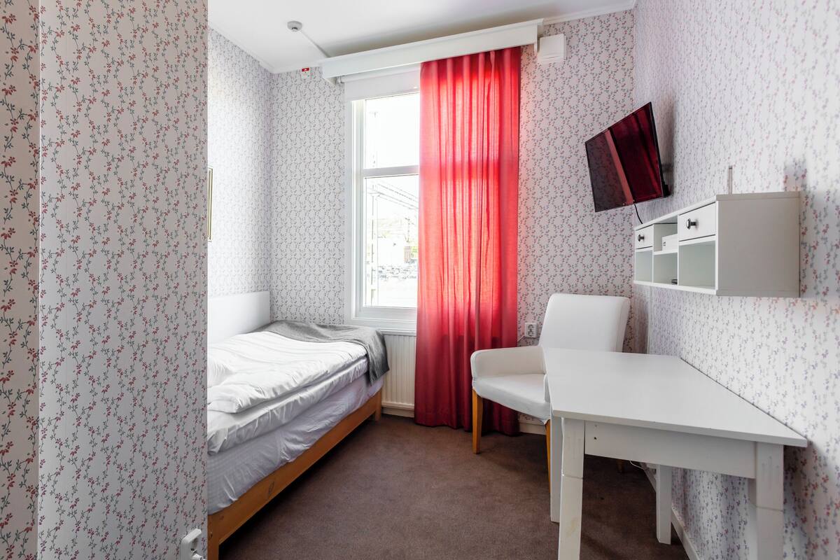 Ein Schlafzimmer mit einem Einzelbett, einem Schreibtisch, einem Stuhl, einem Fernseher und einem Fenster mit roten Vorhängen.