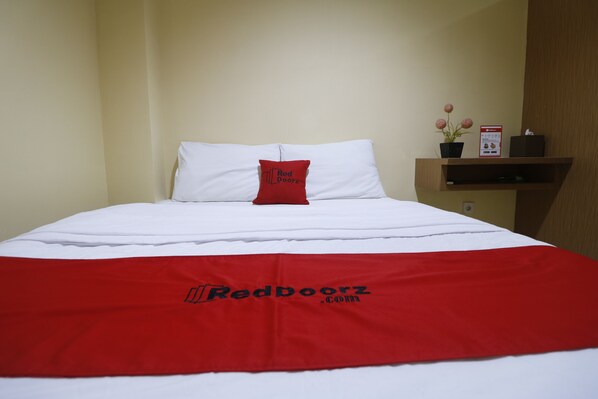 Una cama bien hecha con una colcha blanca y roja, una almohada roja, y una pequeña estantería con una planta en maceta y un letrero.