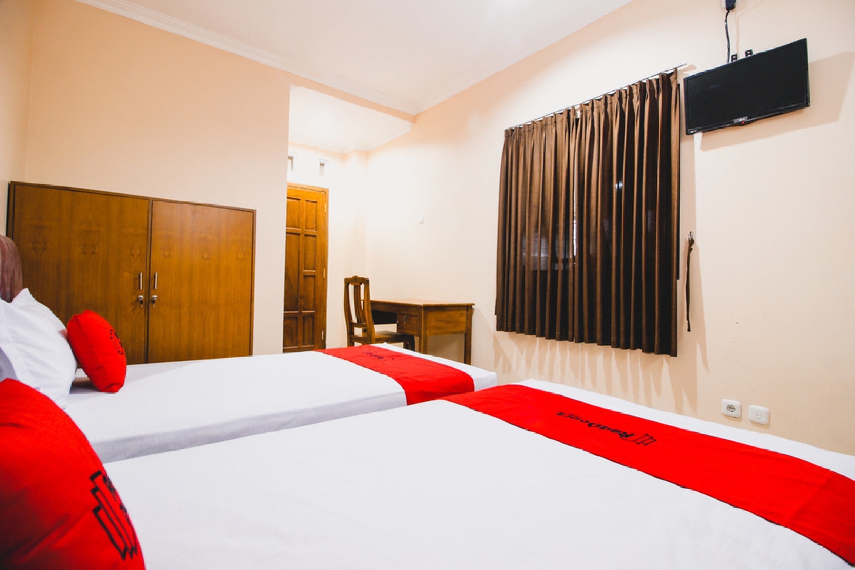 A hotel room with two beds, a wooden wardrobe, a desk, and a TV mounted on the wall.