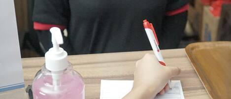 Une personne est assise à un bureau et remplit un formulaire. On peut voir à côté d’elle un flacon rose de gel hydroalcoolique et un stylo blanc.