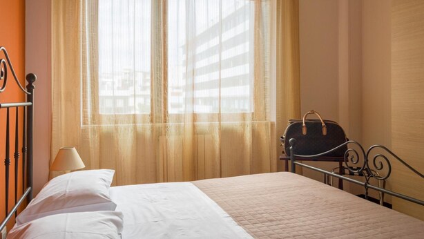 Een opgemaakt bed met een gewatteerde spreideken, een bedlamp en een handtas op een metalen bedframe.