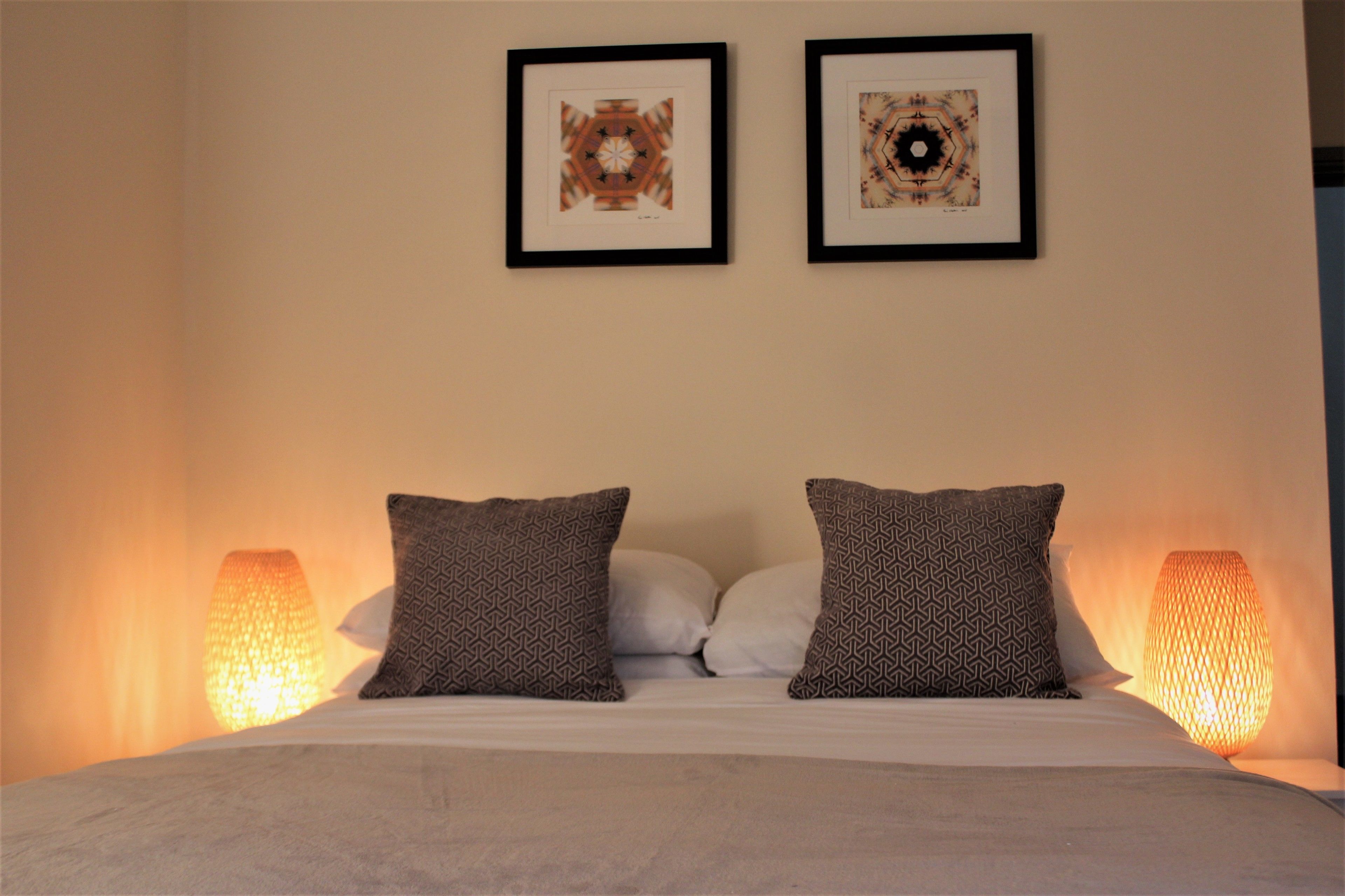 Una cama impecablemente arreglada con almohadones estampados, dos cuadros enmarcados en la pared y dos lámparas fijadas a la pared.