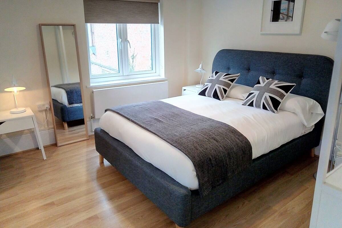 A bedroom with a large bed, a bedside table with a lamp, a window with a view of the outside, and a framed picture on the wall.