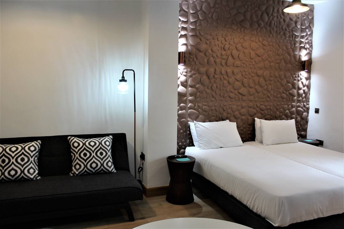 A modern hotel room with a bed, a sofa, a lamp, and a textured wall.
