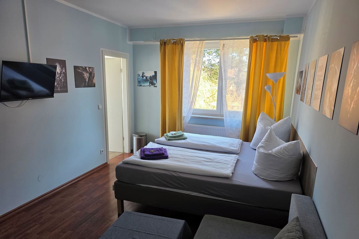 A room with a bed, a small table, a TV mounted on the wall, and a window with curtains.