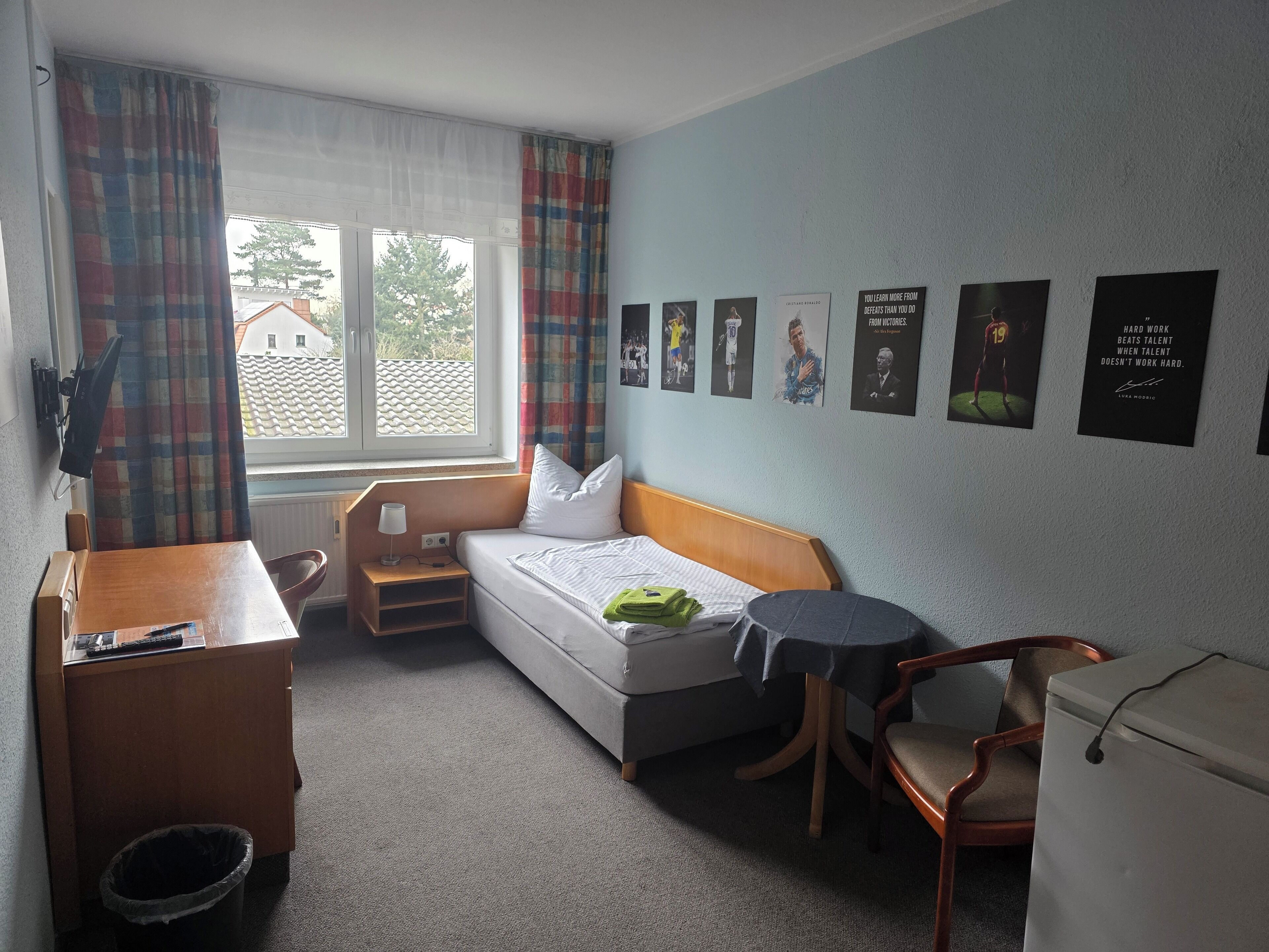 Ein kleines Hotelzimmer mit Bett, Schreibtisch, Stuhl und einem kleinen Tisch. Vor dem Fenster hängen Vorhänge und an der Wand befinden sich mehrere gerahmte Bilder.