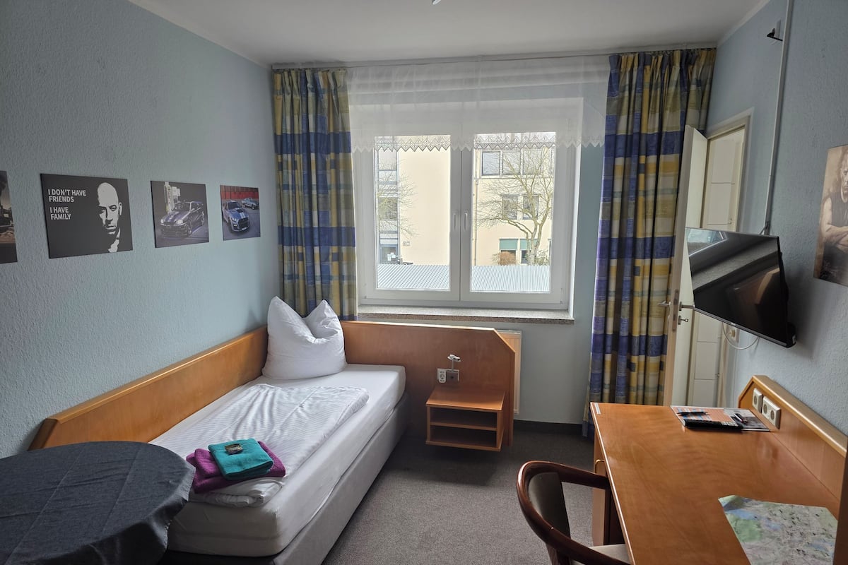 A hotel room with a single bed, a small bedside table, a desk with a chair, a TV, and a window with curtains.