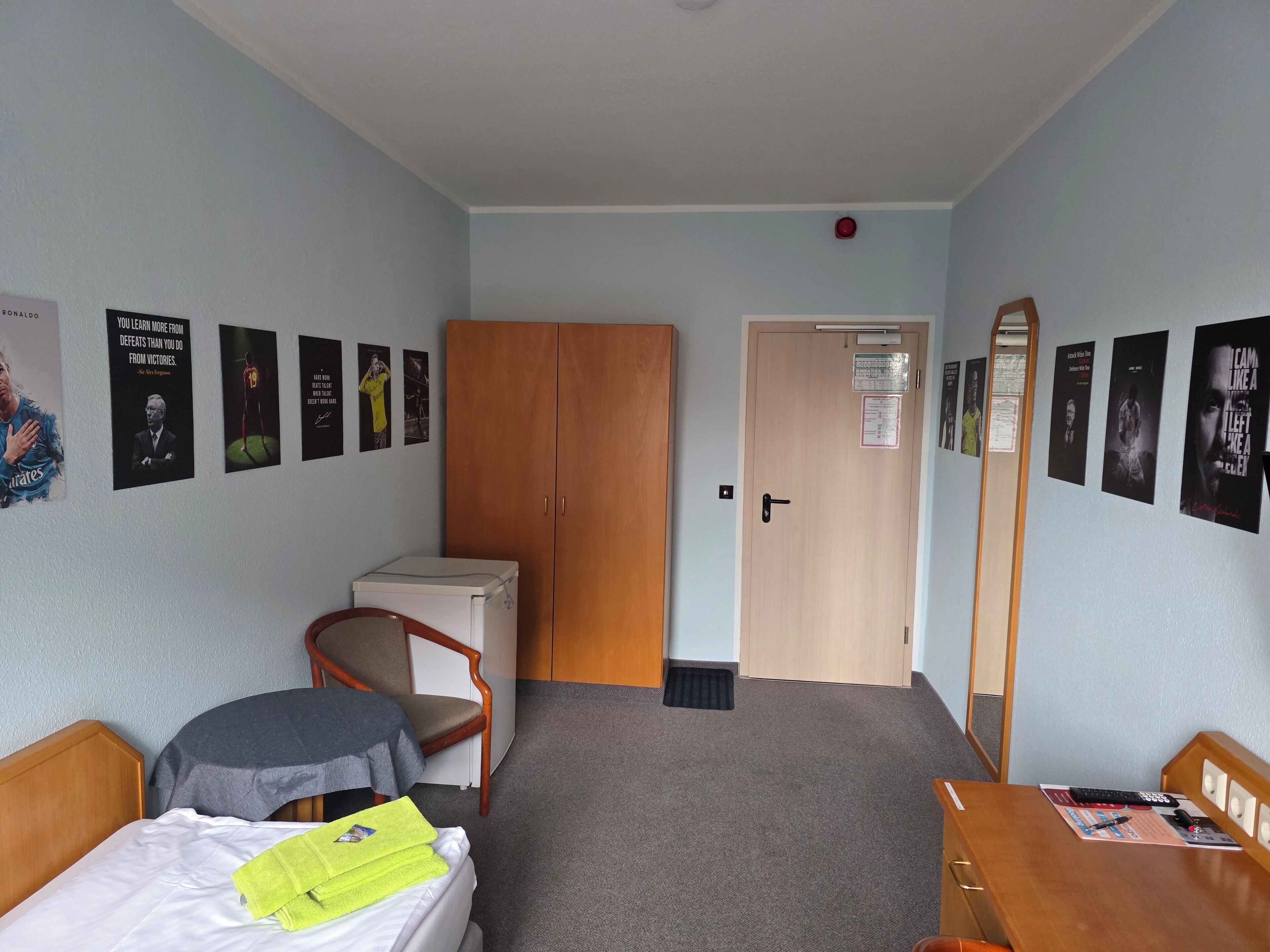 Habitación de hotel con cama, escritorio, armario y puerta. Hay carteles en las paredes y un detector de incendios rojo en el techo.