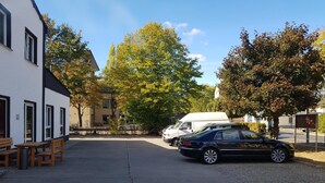 Un aparcamiento con un coche negro y una furgoneta blanca, un edificio de fachada blanca y detalles negros, y árboles con follaje otoñal.