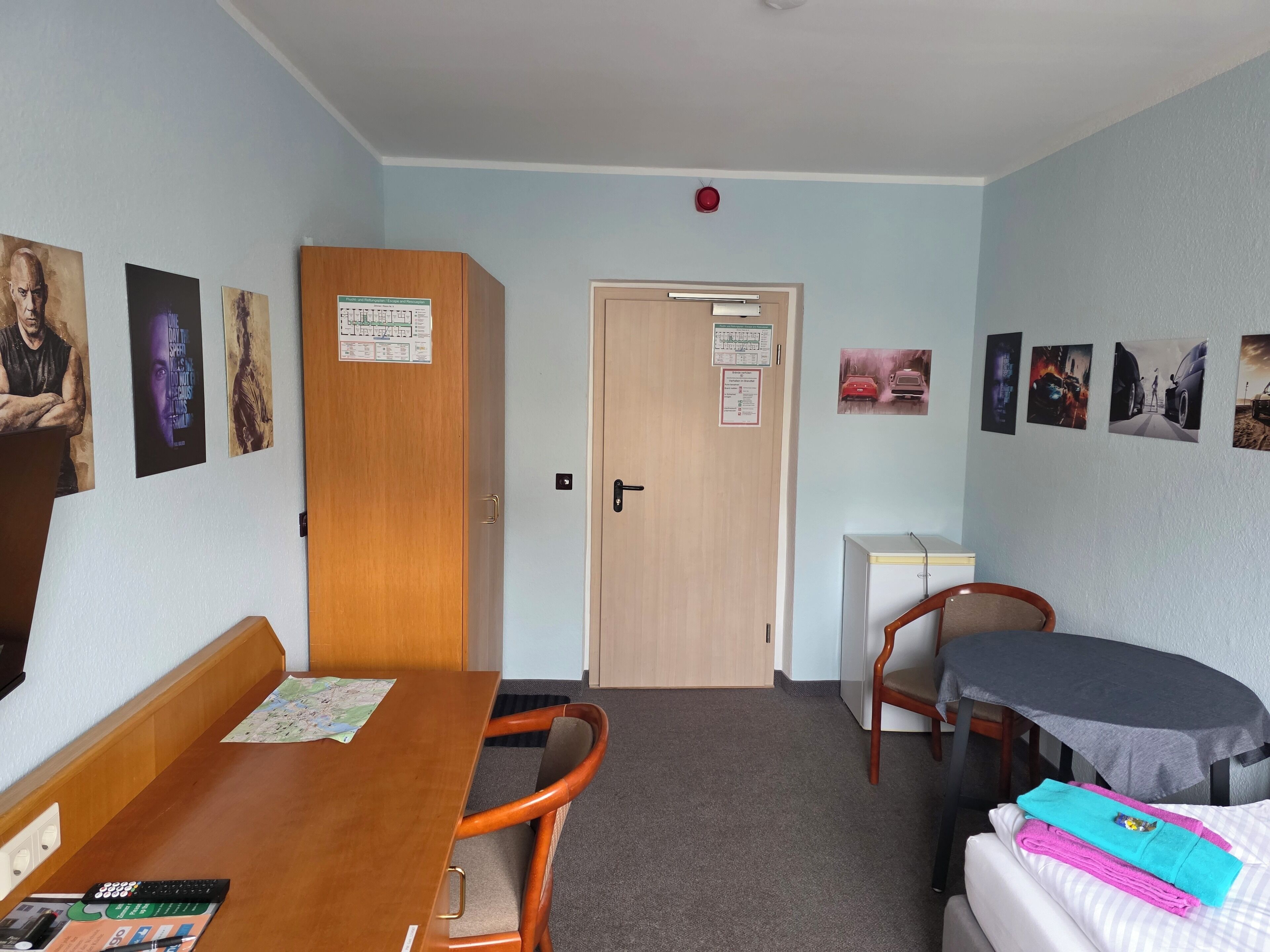 A hotel room with a wooden desk, a bed with a gray blanket, a small table, and a chair. There are posters on the walls and a door with a sign.