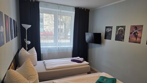 Habitación de hotel con dos camas, un ventanal grande con cortinas, un televisor de pantalla plana y varias fotografías enmarcadas en la pared.