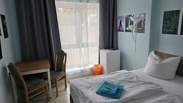 A hotel room with a bed, a desk, a chair, a window with curtains, and a wall with framed pictures.
