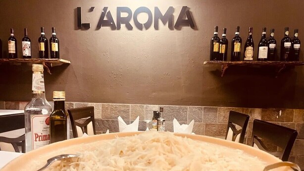 Interior de restaurante con un plato grande de queso rallado en primer plano, una pared con la señalización 'L'AROMA' y estantes con botellas de vino.