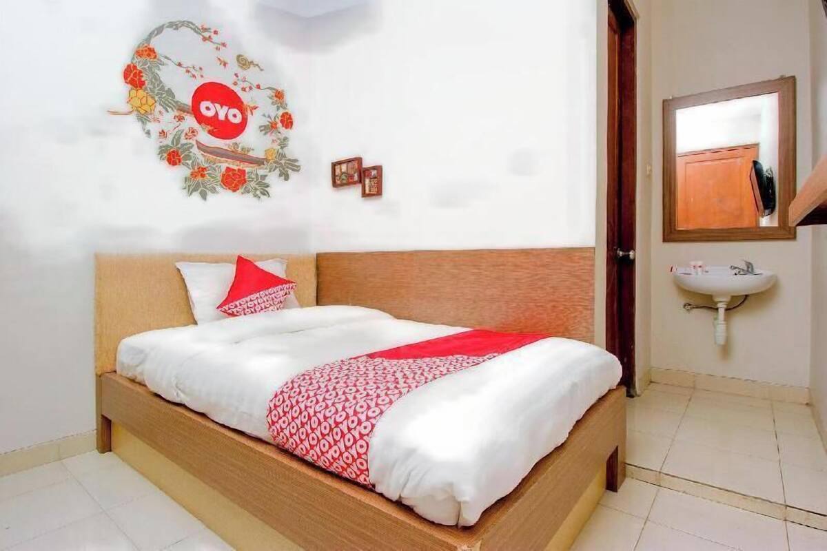 Un lit avec une tête de lit en bois, une couvre-lit blanche et rouge, un oreiller rouge, une décoration murale avec l’inscription « OYO » et un motif floral, ainsi qu’un lavabo blanc avec un miroir au-dessus.
