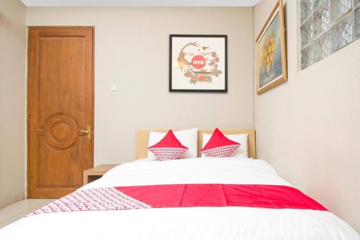Une chambre avec une porte en bois, un lit avec une literie blanche et rouge, et un tableau encadré au mur.