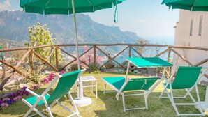 Outdoor seating with green cushions and white frames, surrounded by a wooden fence and flowering plants, with a mountain view in the background. - Mimì B&B (Ravello)