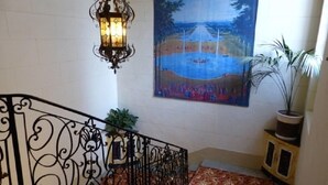 A staircase with ornate black metal railings, a decorative wall-mounted light fixture, and a large landscape painting above. - Hotel Du Fiacre (Carpentras)