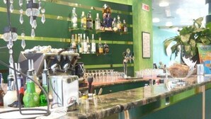 Angolo bar con parete verde, scaffali pieni di bottiglie, macchina per caffè e una pianta.