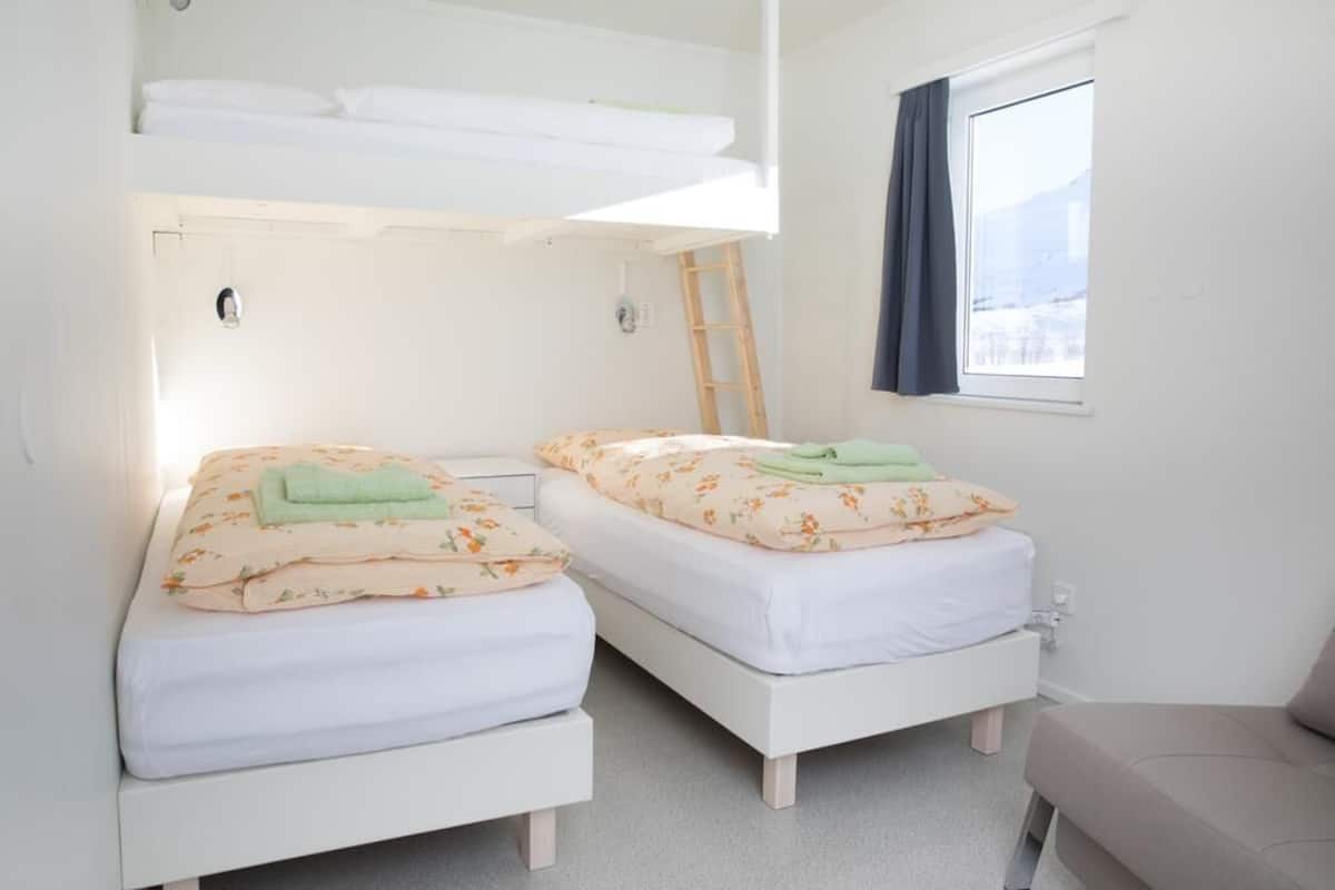 Een kamer met twee bedden, een stapelbed bovenin, een raam met gordijnen en een stoel.