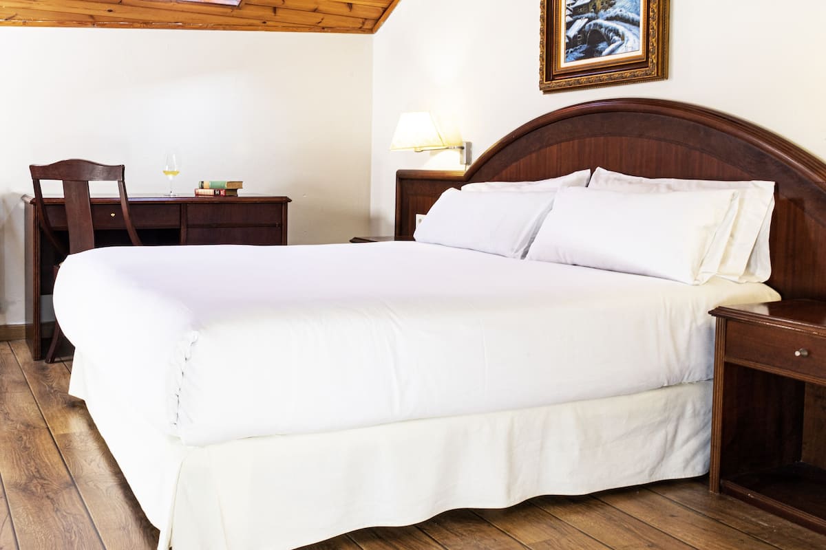 A bedroom with a wooden ceiling, a bed with white linens, a desk with books, a chair, and a framed picture on the wall.