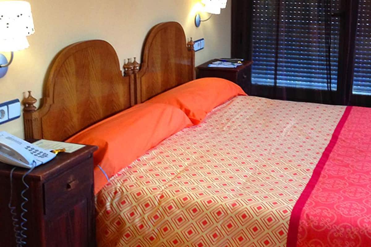 A hotel room with a wooden headboard, a telephone on a nightstand, a patterned bedspread, and a window with blinds.