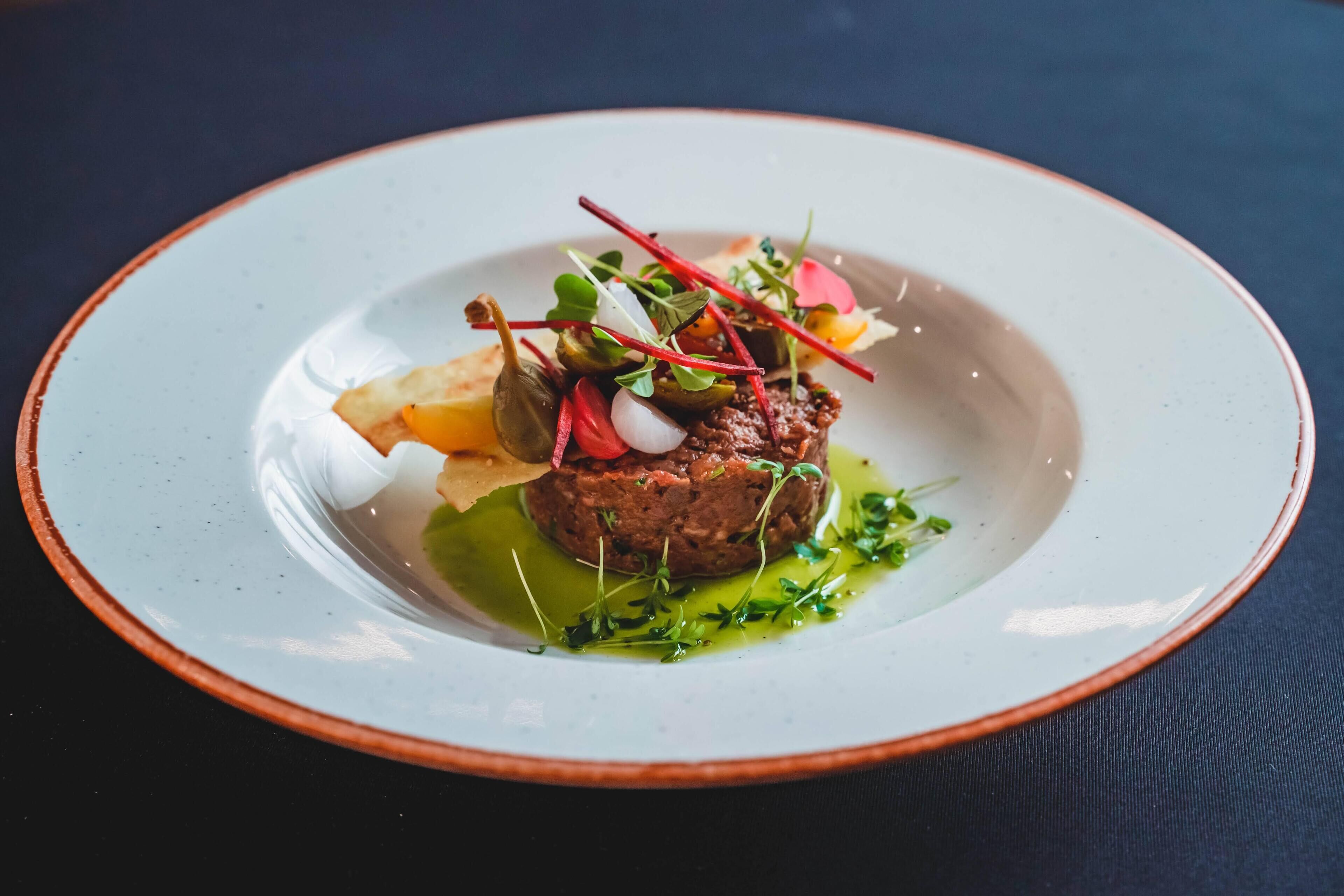 A plate of food with a central meat patty, garnished with greens and vegetables, on a white plate with a brown rim.