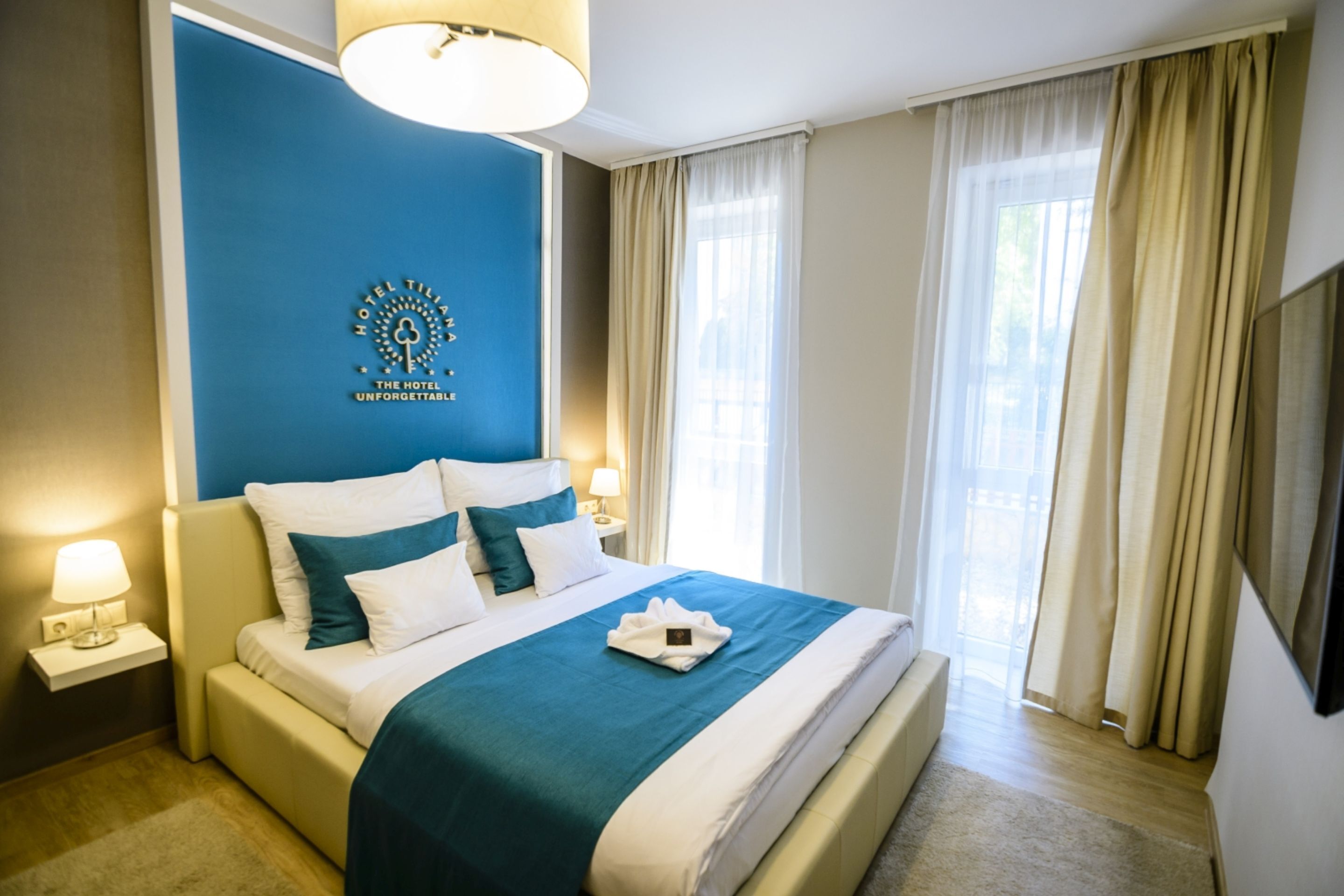 A hotel room with a large bed, a blue accent wall, a flat-screen TV, and a window with curtains.