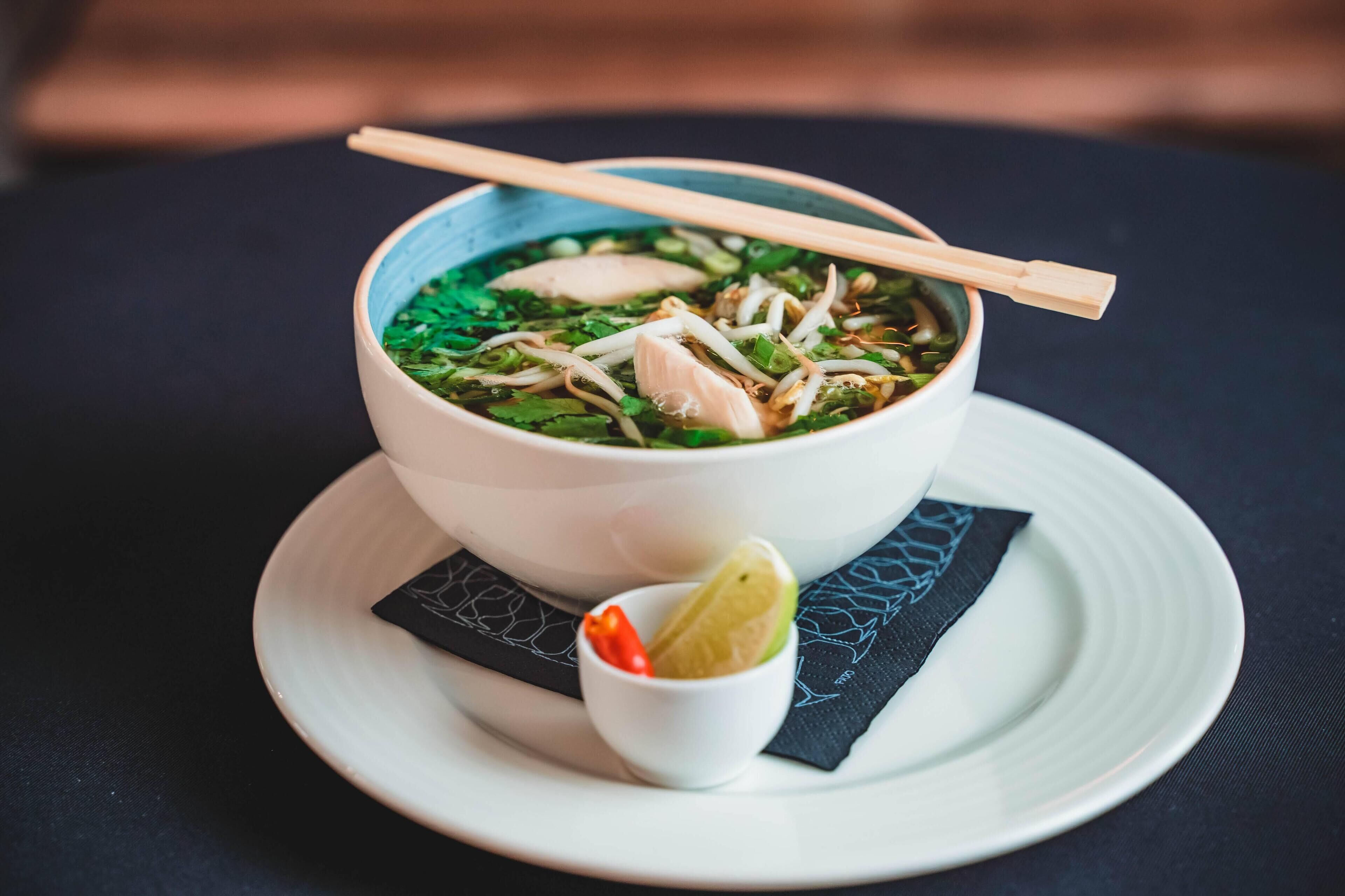 A bowl of soup with chopsticks, garnished with herbs and bean sprouts, served on a white plate with a black napkin underneath.