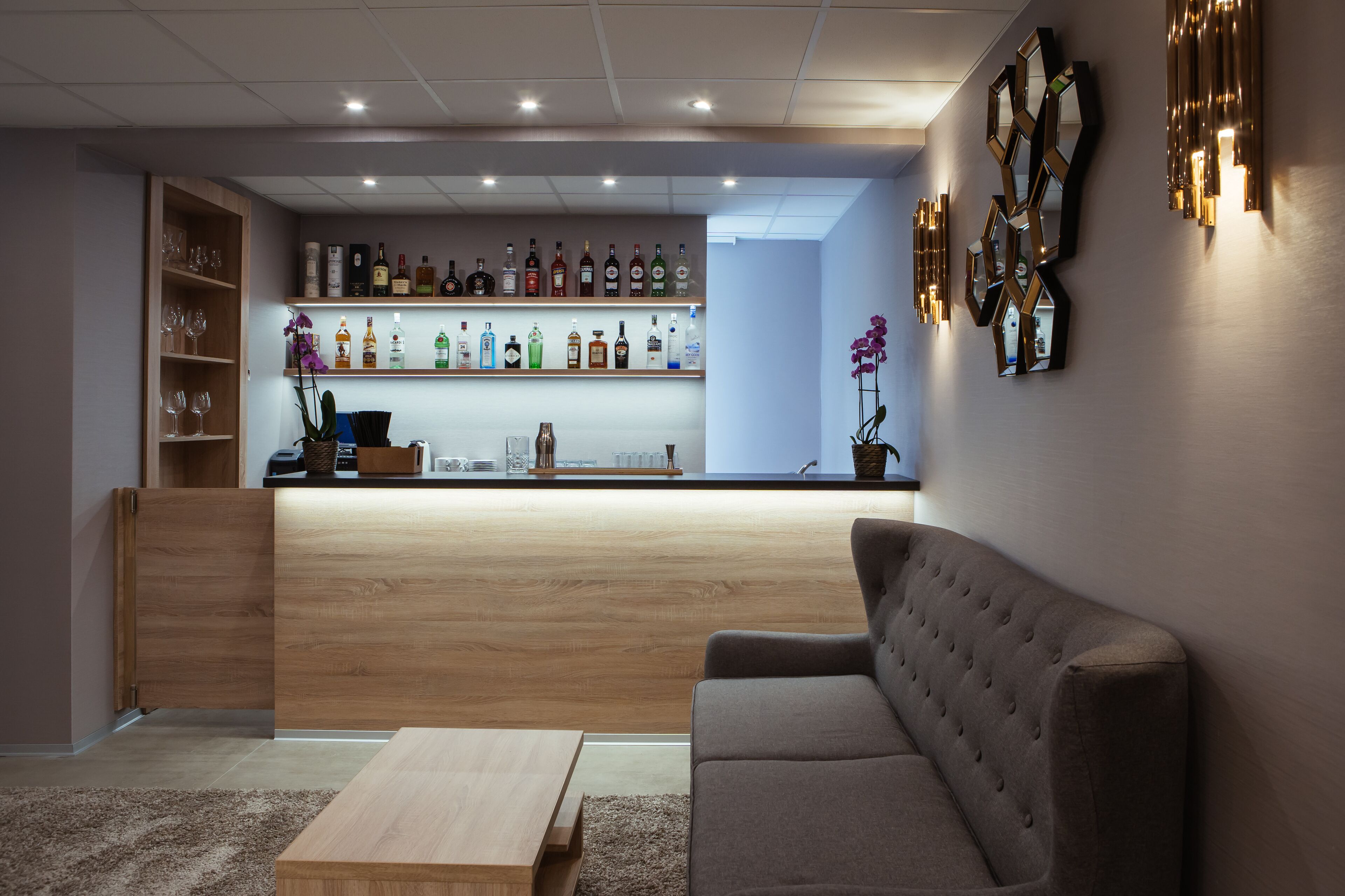 Zona de bar moderna amb prestatgeries de fusta que mostren diferents ampolles i gots, una zona d'seients amb seients còmodes i una taula de café, i aplics decoratius a la paret.