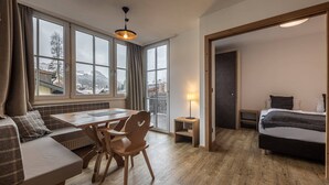 Habitación de hotel moderna con piso de madera, un comedor con mesa y sillas, una cama con cabecera oscura y ventanas amplias que ofrecen vistas al exterior.