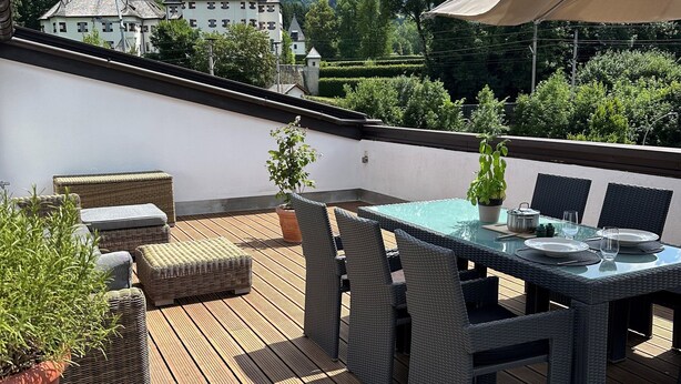 Una terraza de madera con mobiliario de mimbre y una mesa lista para comer, con vistas a un paisaje montañoso.