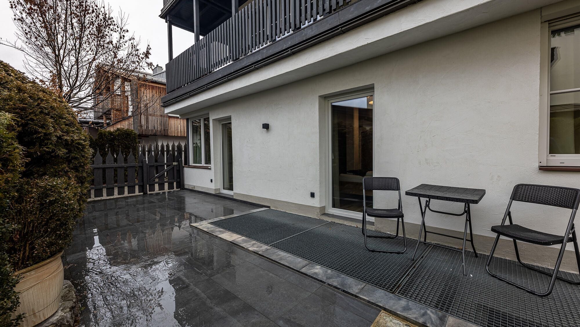 A modern building with a balcony, a small outdoor seating area with two chairs and a table, and a gated entrance.