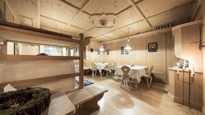 A cozy room with wooden walls and ceiling, featuring a dining area with tables and chairs, a bench, and a small altar with candles. - Alpenhof (Ultimo)