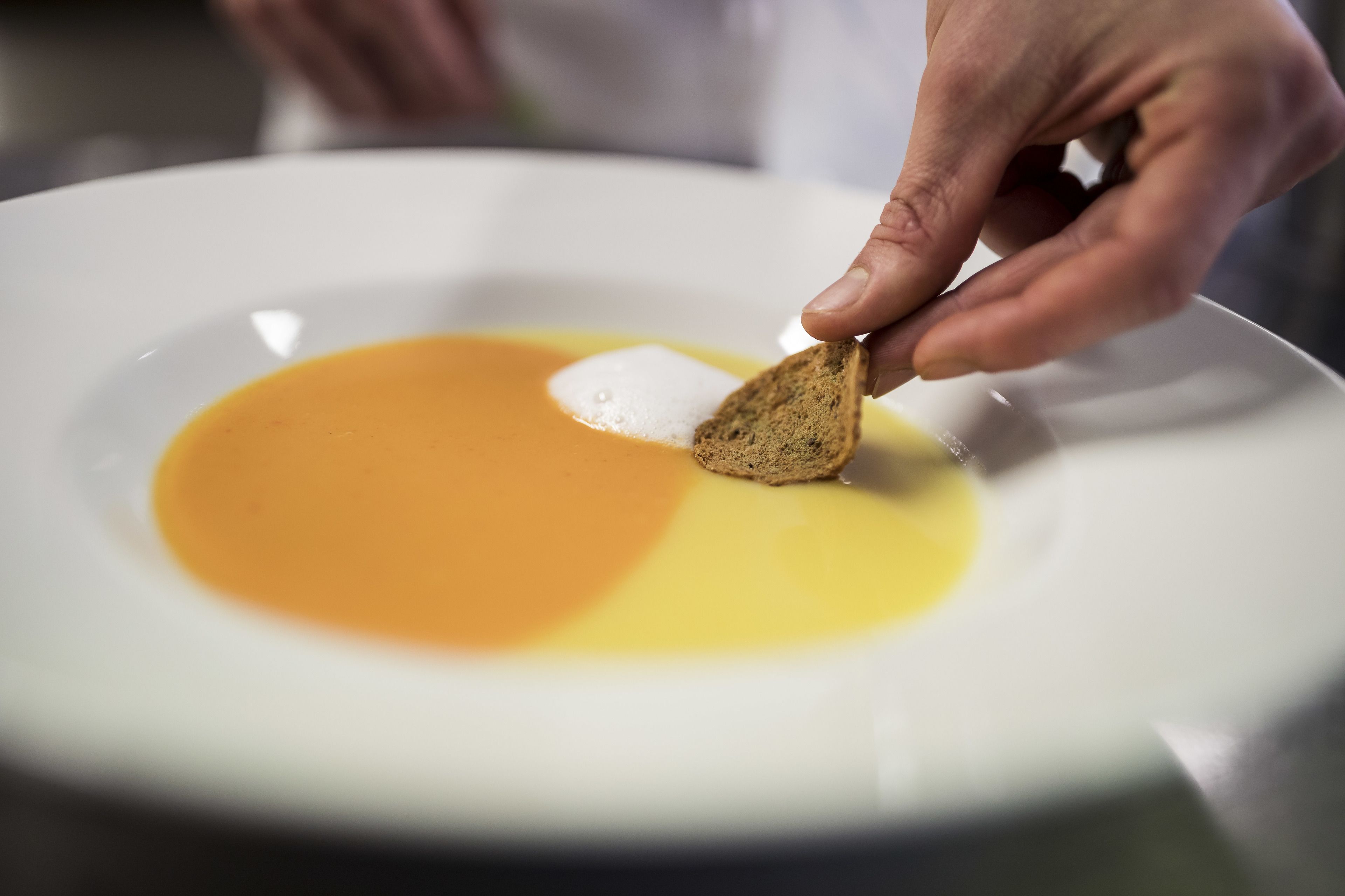 Een hand legt een sneetje toast neer op een bord met een lepel slagroom en een plas vloeistof.