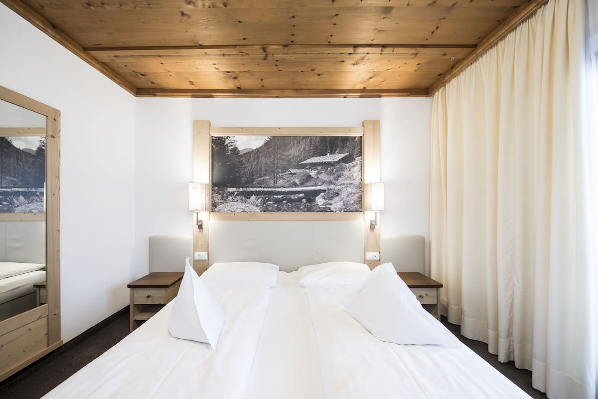Habitación de hotel con techo de madera, cama con ropa blanca, mesita de noche, espejo y un cuadro de un paisaje montañoso.