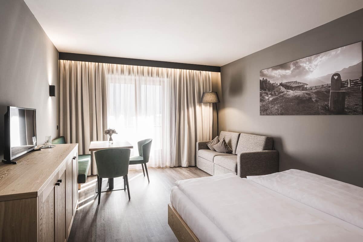 Habitación de hotel con cama, escritorio con computadora, silla, sofá y ventana grande con cortinas.