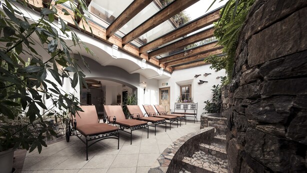 A sunlit indoor patio with stone-tiled flooring, wooden beams, and a stone wall. - Alpenhof (Ultimo)