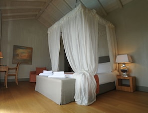 A bedroom with a four-poster canopy bed, a bedside table with a lamp, a chair, and a painting on the wall. - Paparouna (Alonissos)