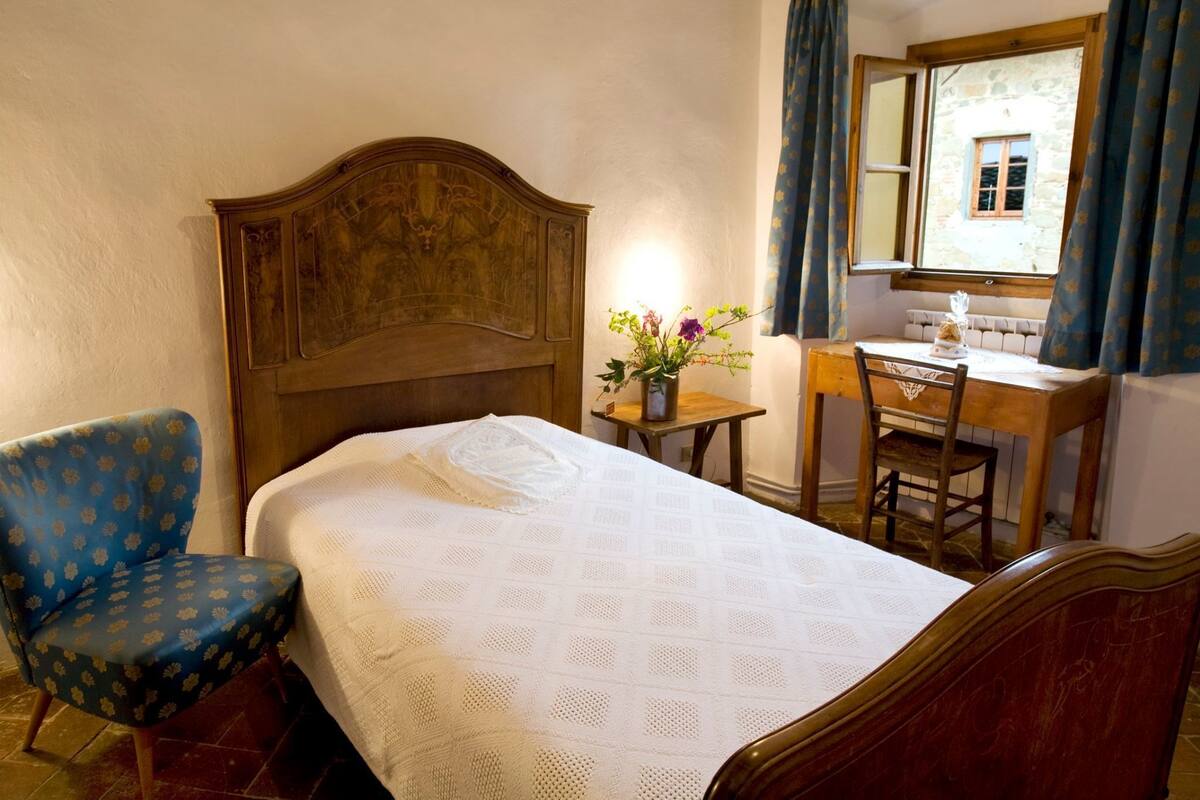 Ein Schlafzimmer mit hölzernem Kopfteil, einem Einzelbett mit weißer Bettdecke, einem blauen Sessel, einem Holztisch mit Stuhl und einem Fenster mit Vorhängen.
