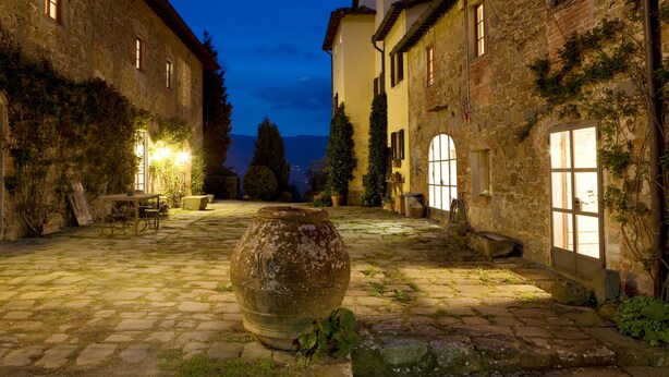 Une cour en pierre avec un grand vase, un chemin pavé et des bâtiments couverts de lierre et dotés de fenêtres.