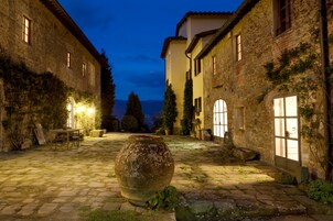 Un patio de piedra con un jarrón grande, un camino empedrado y edificios con hiedra y ventanas.