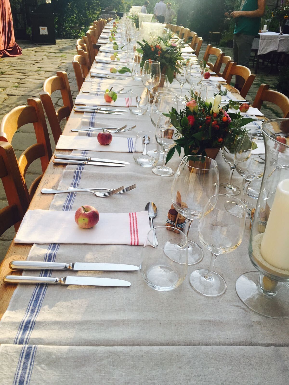 Une longue table à manger, dressée avec des chaises, des assiettes, des couverts et des verres à vin, agrémentée de fleurs décoratives et de pommes.
