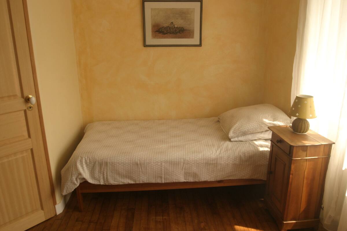 Ein kleines Zimmer mit einem Einzelbett, Holzboden, einem Nachttisch mit Lampe und einem gerahmten Bild an der Wand.
