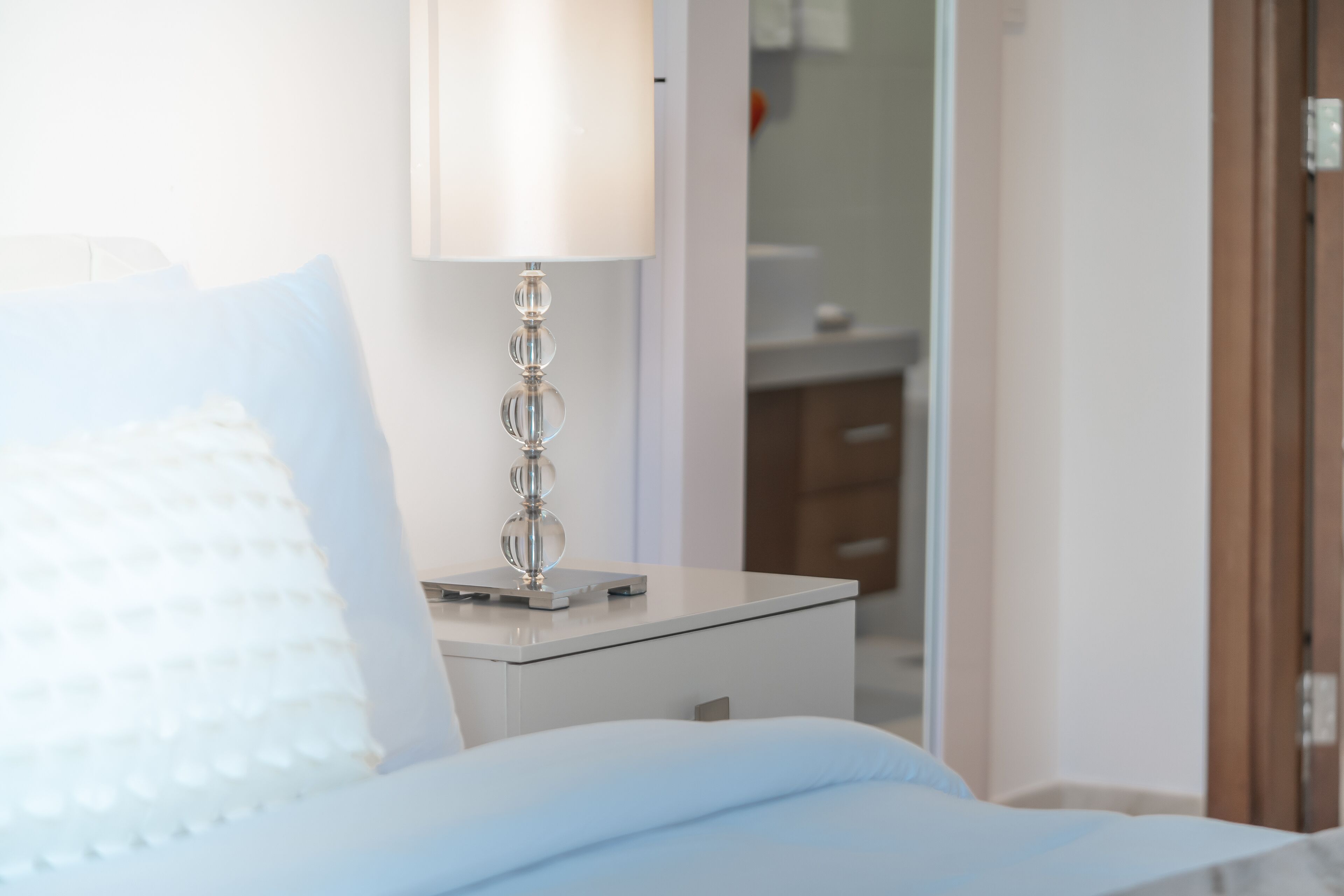 A neatly made bed with a white pillow and a white duvet, a bedside table with a lamp, and a wooden door in the background.