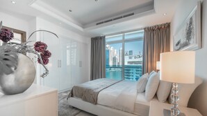 Een moderne slaapkamer met een groot bed, een nachtkastje met een lamp, een decoratieve vaas, en uitzicht op de stad door grote ramen.