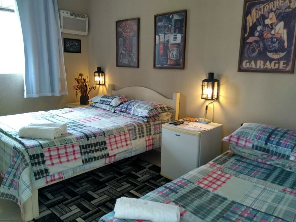 Quarto de hotel com duas camas, cada uma com colcha xadrez e toalha branca. Há unidades de ar condicionado montadas na parede e cartazes emoldurados.